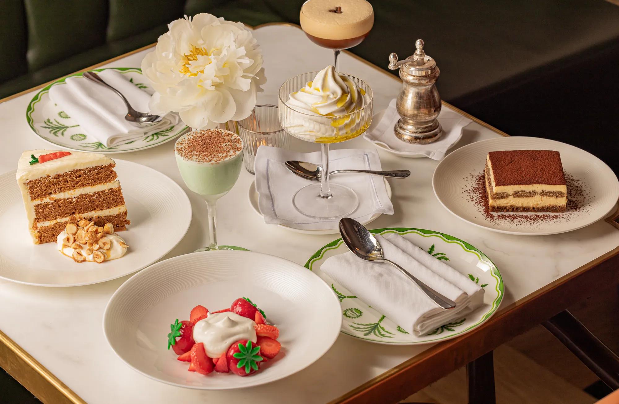 Dessert selection including carrot cake, tiramisu, strawberries, cocktails, and whipped cream on a marble table.