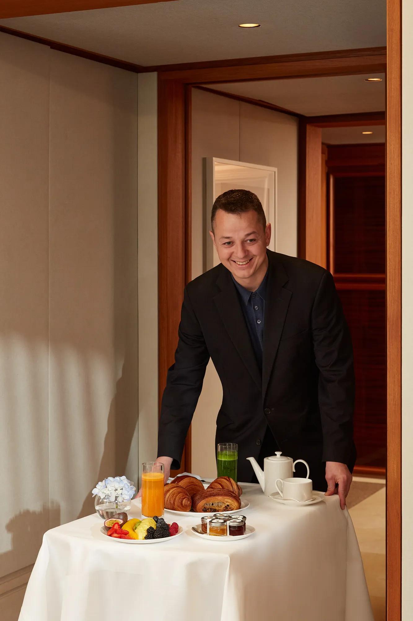 Smiling server presenting table with croissants, fresh fruit, juices, jams, and tea in hotel suite.