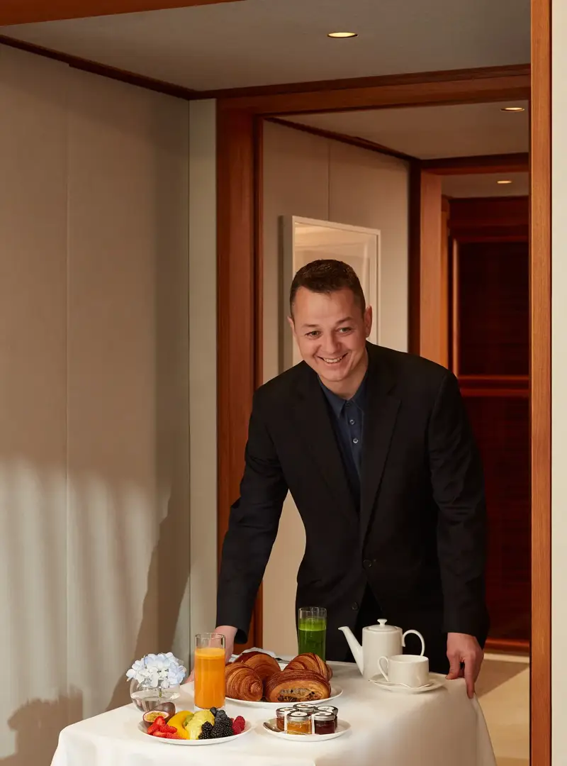 Smiling server presenting table with croissants, fresh fruit, juices, jams, and tea in hotel suite.