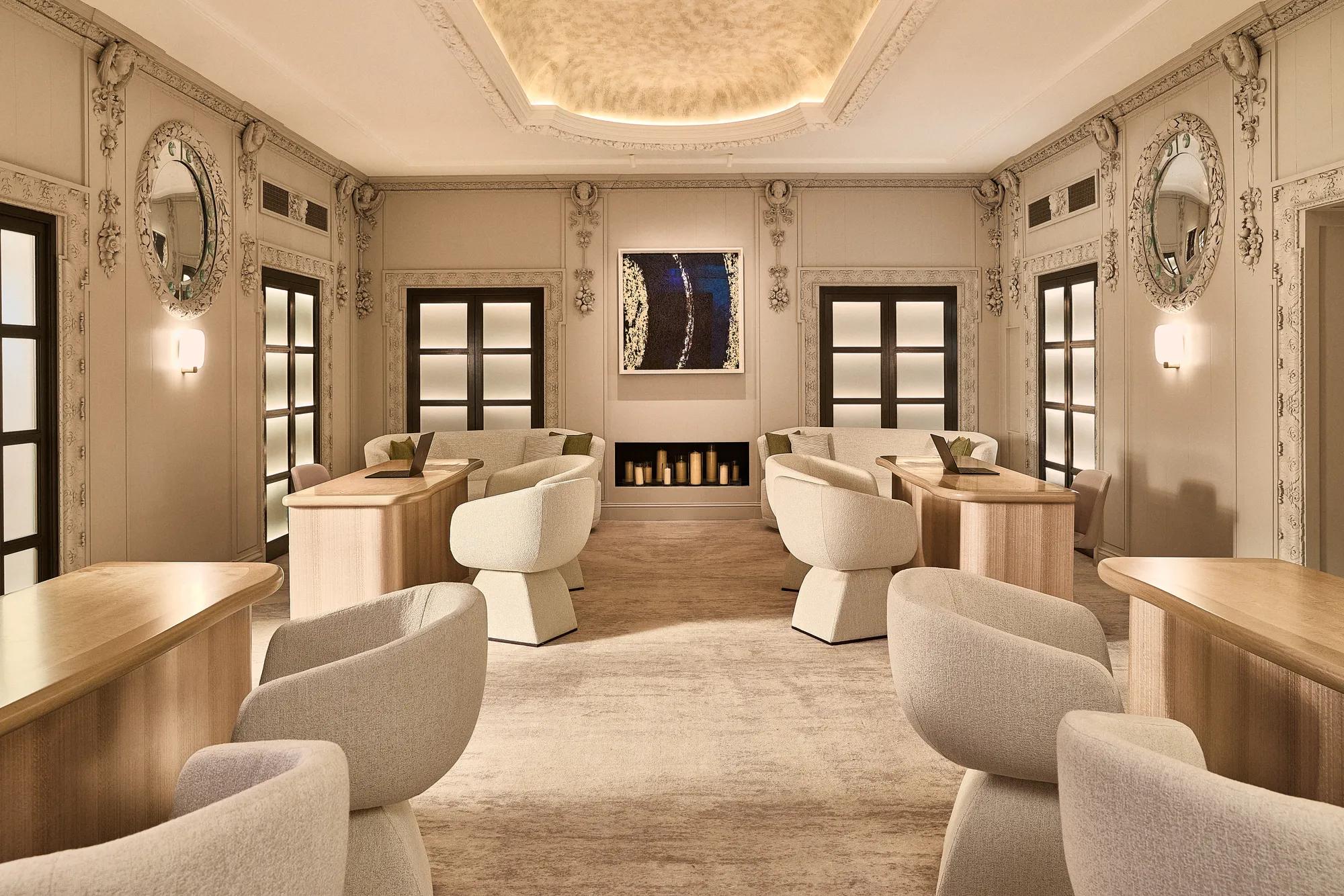 A softly lit lounge with cream armchairs, light wood tables and ornate wall detailing, facing a fireplace and contemporary artwork.