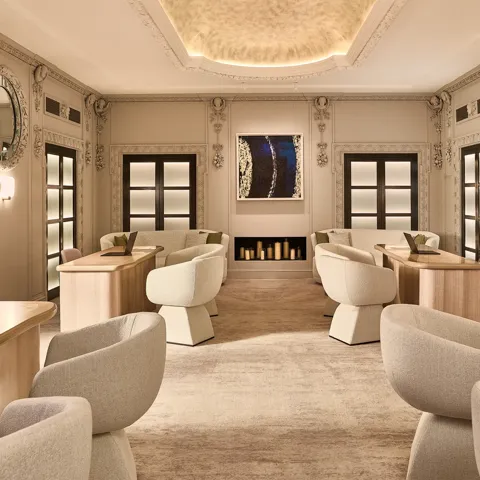 A softly lit lounge with cream armchairs, light wood tables and ornate wall detailing, facing a fireplace and contemporary artwork.