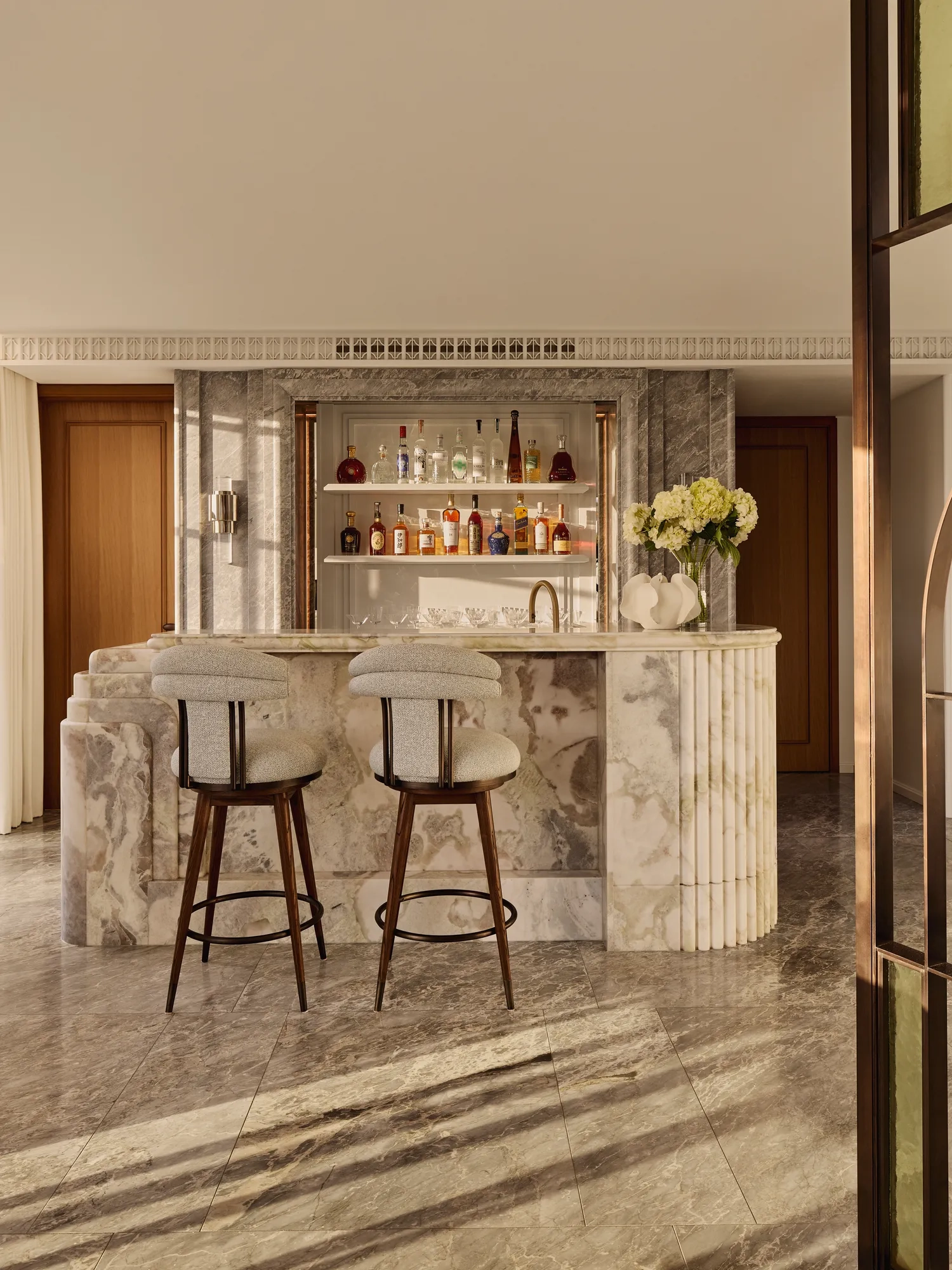 Sophisticated marble bar area with two upholstered stools, backlit shelves displaying assorted spirits, and a vase of white flowers, set against warm wood and stone finishes.