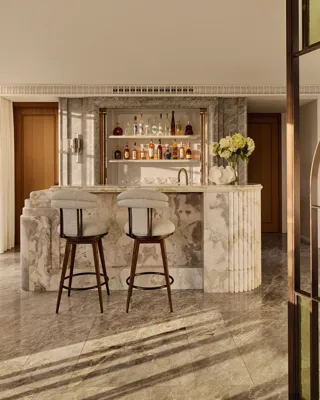 Sophisticated marble bar area with two upholstered stools, backlit shelves displaying assorted spirits, and a vase of white flowers, set against warm wood and stone finishes.