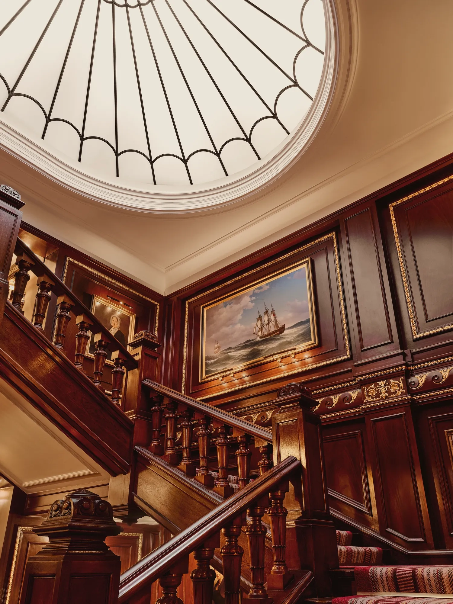 Grand wooden staircase with ornate paneling, nautical painting, and large domed skylight above.