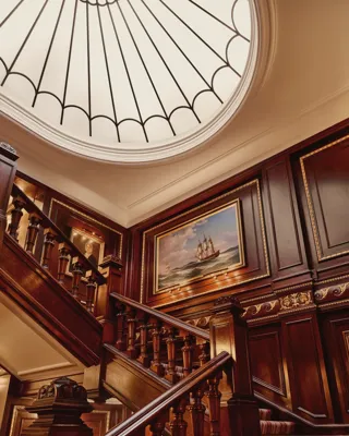 Grand wooden staircase with ornate paneling, nautical painting, and large domed skylight above.