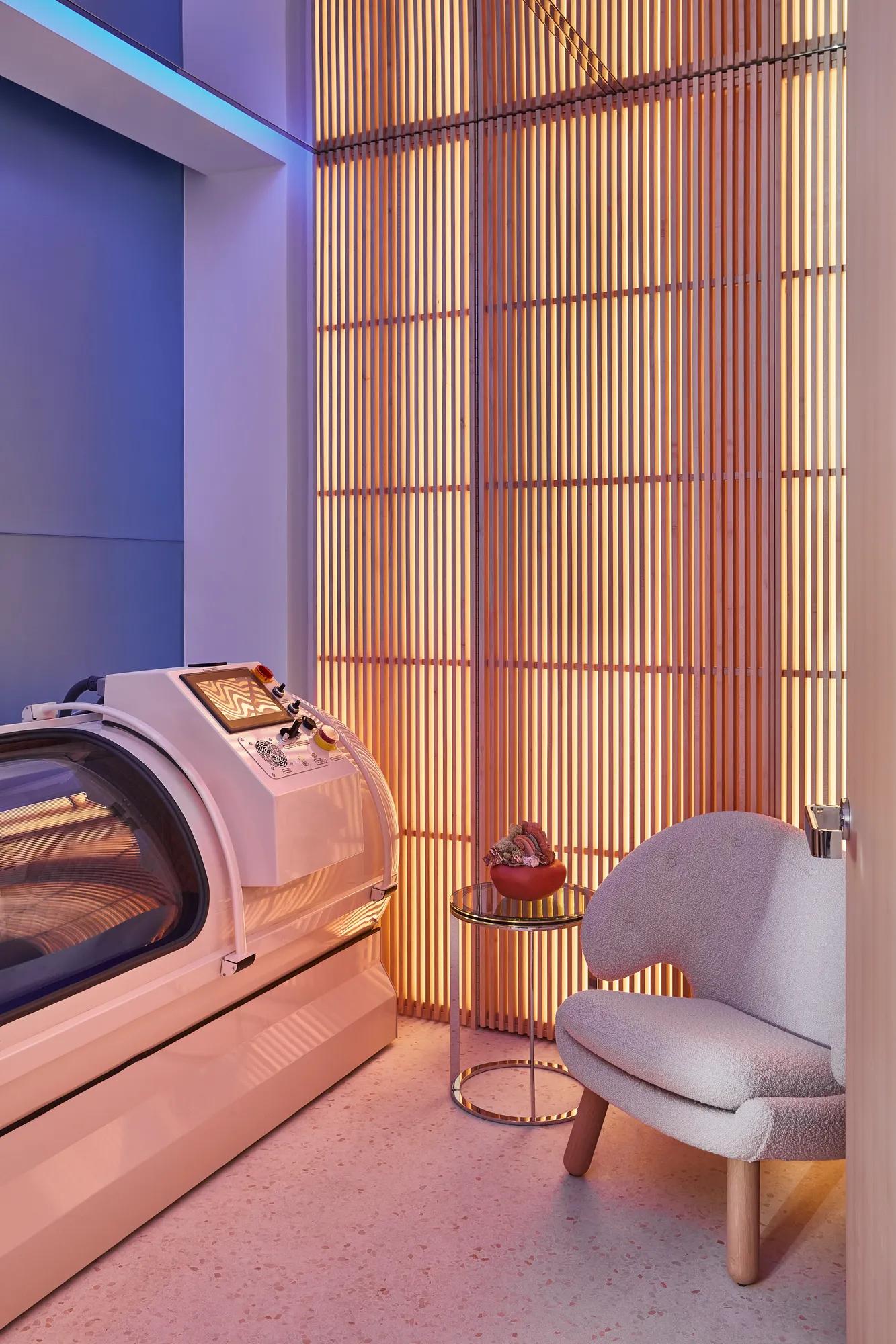 Modern wellness room with glowing slatted wall, sleek white hyperbaric chamber, small side table, and cosy upholstered armchair.