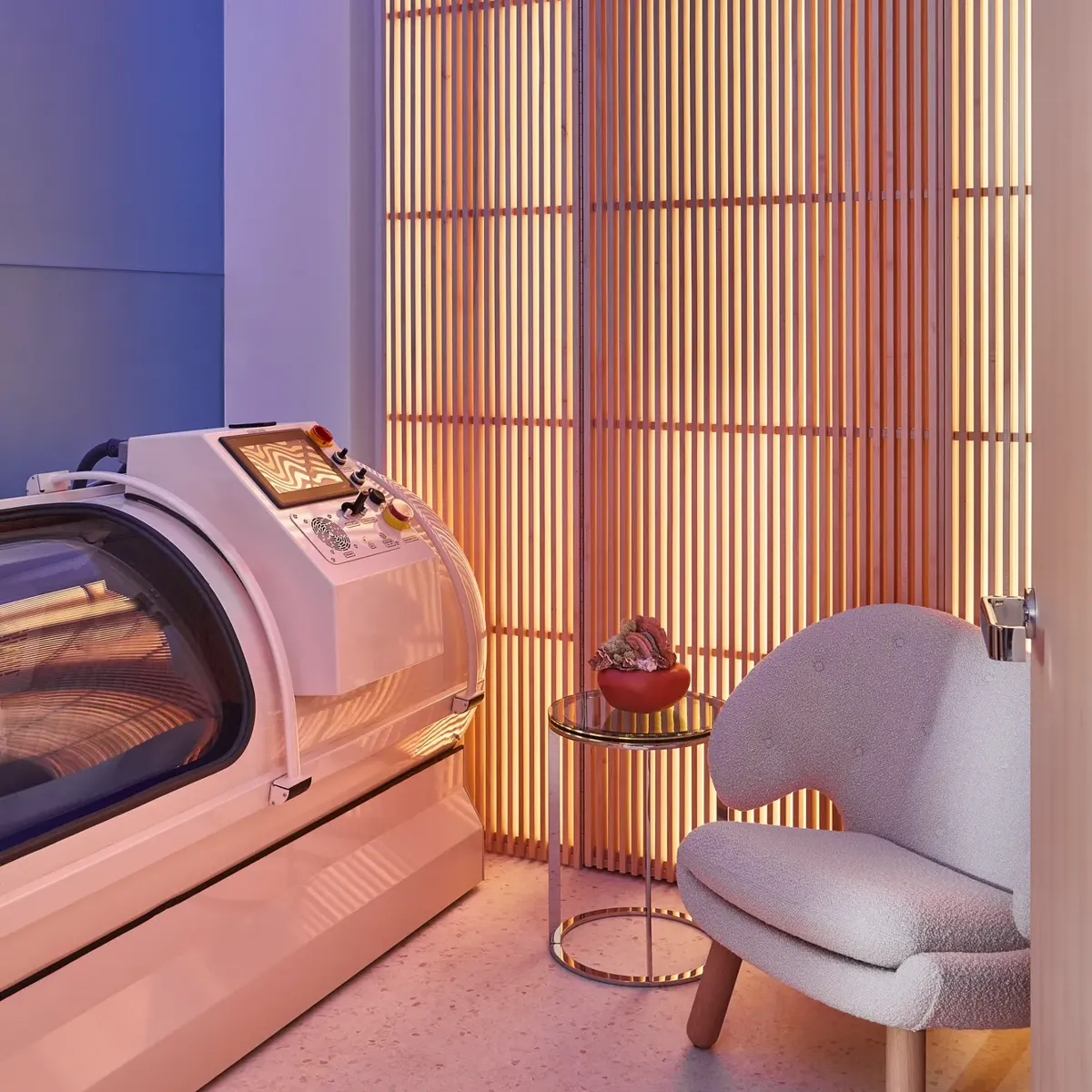 Modern wellness room with glowing slatted wall, sleek white hyperbaric chamber, small side table, and cosy upholstered armchair.