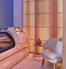 Modern wellness room with glowing slatted wall, sleek white hyperbaric chamber, small side table, and cosy upholstered armchair.