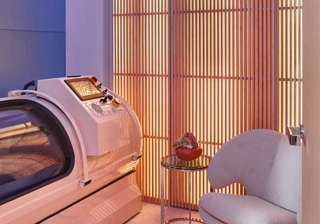 Modern wellness room with glowing slatted wall, sleek white hyperbaric chamber, small side table, and cosy upholstered armchair.