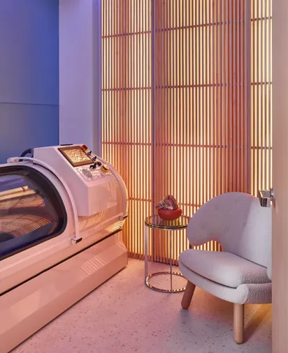 Modern wellness room with glowing slatted wall, sleek white hyperbaric chamber, small side table, and cosy upholstered armchair.