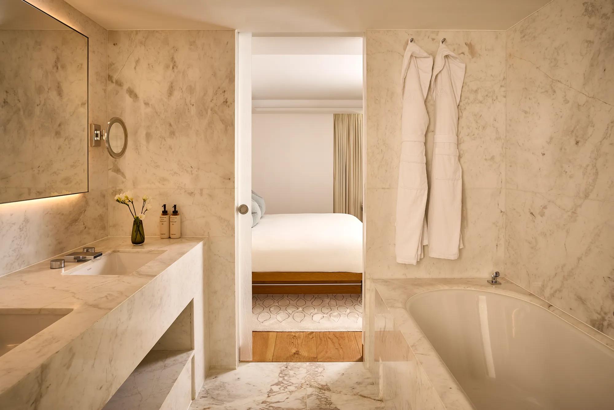 Marble bathroom with a soaking bath, vanity, illuminated mirror and sliding doorway leading to the adjoining bedroom.
