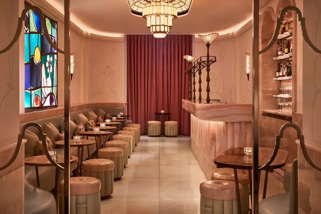 Art Deco–inspired cocktail bar with soft pink and cream tones, featuring a marble counter, upholstered stools, and a row of small tables along a cushioned banquette. A colorful stained-glass window and warm lighting create an intimate, elegant atmosphere.