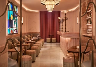 Art Deco–inspired cocktail bar with soft pink and cream tones, featuring a marble counter, upholstered stools, and a row of small tables along a cushioned banquette. A colorful stained-glass window and warm lighting create an intimate, elegant atmosphere.