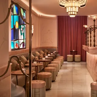 Art Deco–inspired cocktail bar with soft pink and cream tones, featuring a marble counter, upholstered stools, and a row of small tables along a cushioned banquette. A colorful stained-glass window and warm lighting create an intimate, elegant atmosphere.