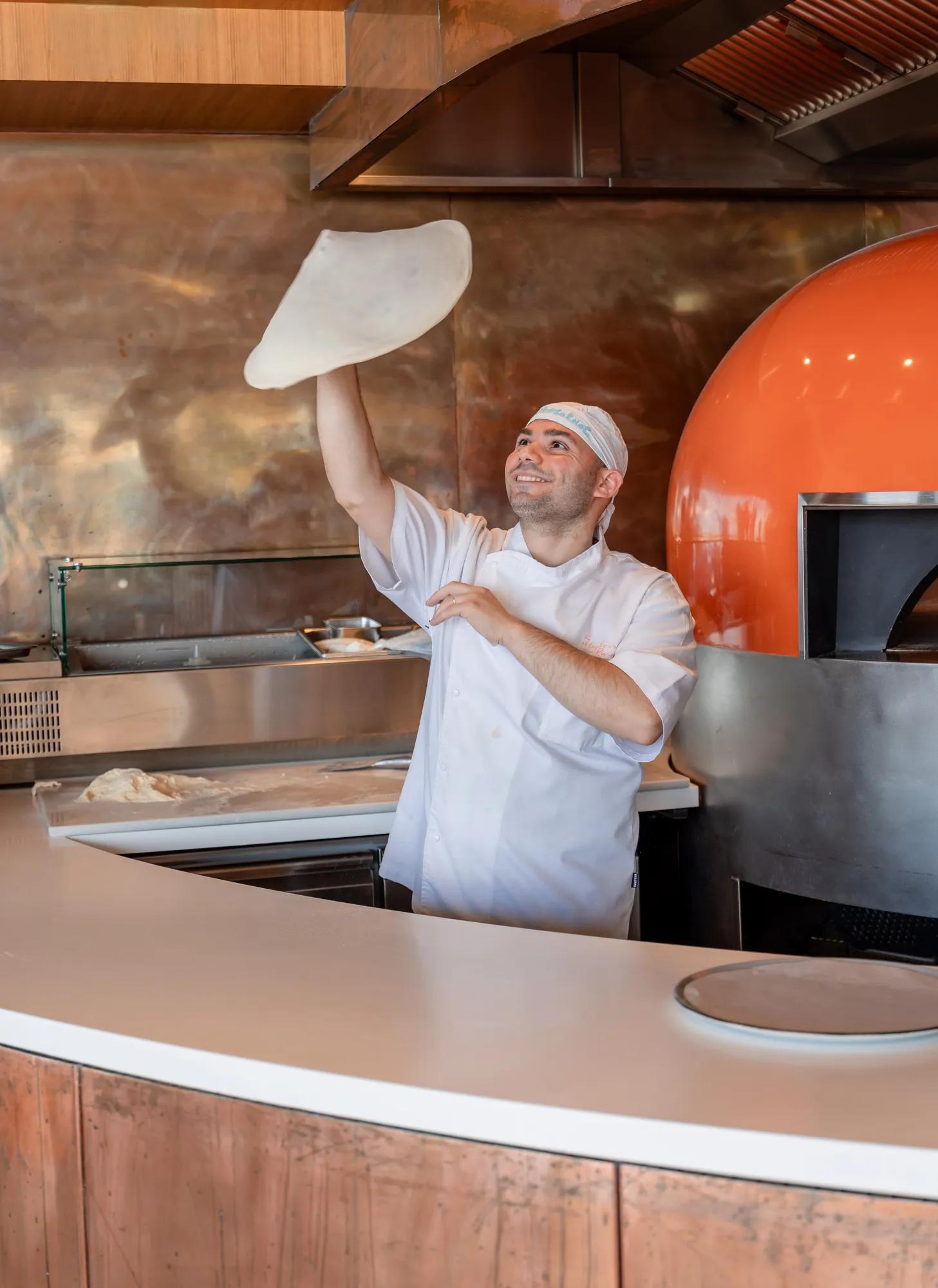 Pizzaiolo lançant une pâte en l’air près d’un four à bois dans une cuisine ouverte.
