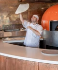 Pizzaiolo lançant une pâte en l’air près d’un four à bois dans une cuisine ouverte.