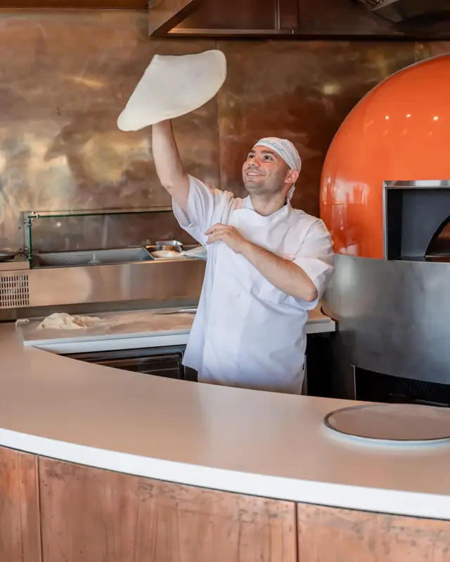 Pizzaiolo lançant une pâte en l’air près d’un four à bois dans une cuisine ouverte.