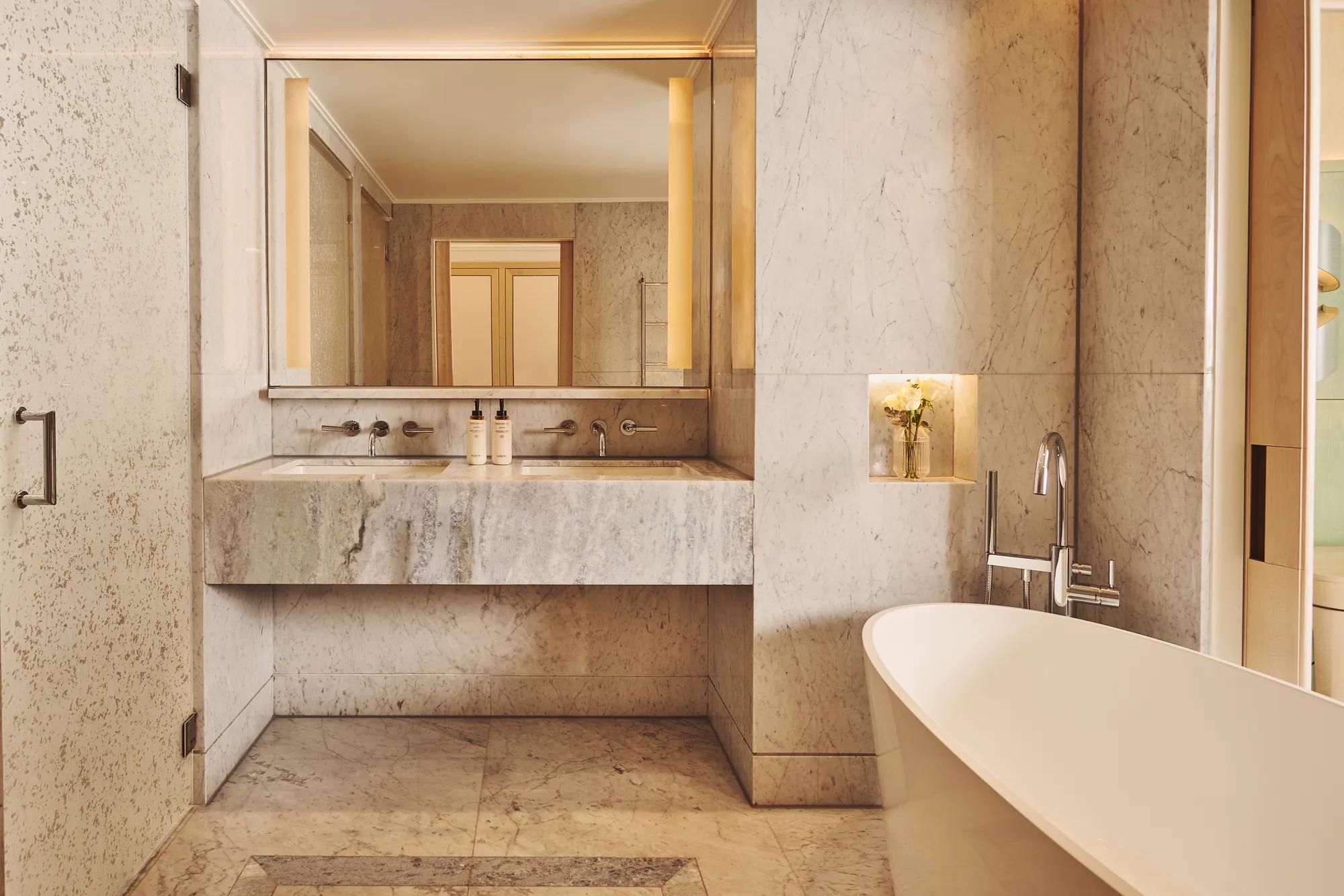 Marble bathroom with freestanding bathtub, vanity with large mirror and lighting, and built-in niche with flowers.