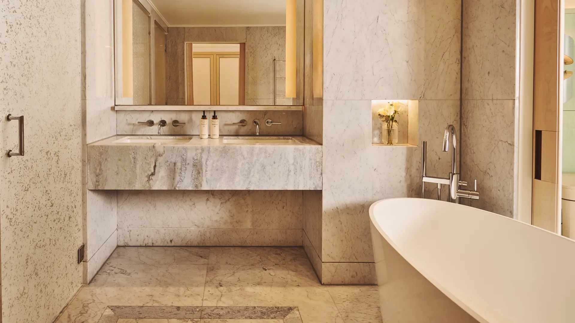 Marble bathroom with freestanding bathtub, vanity with large mirror and lighting, and built-in niche with flowers.