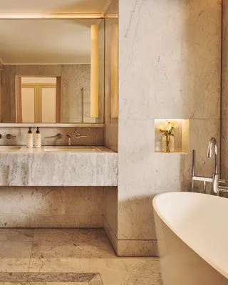 Marble bathroom with freestanding bathtub, vanity with large mirror and lighting, and built-in niche with flowers.