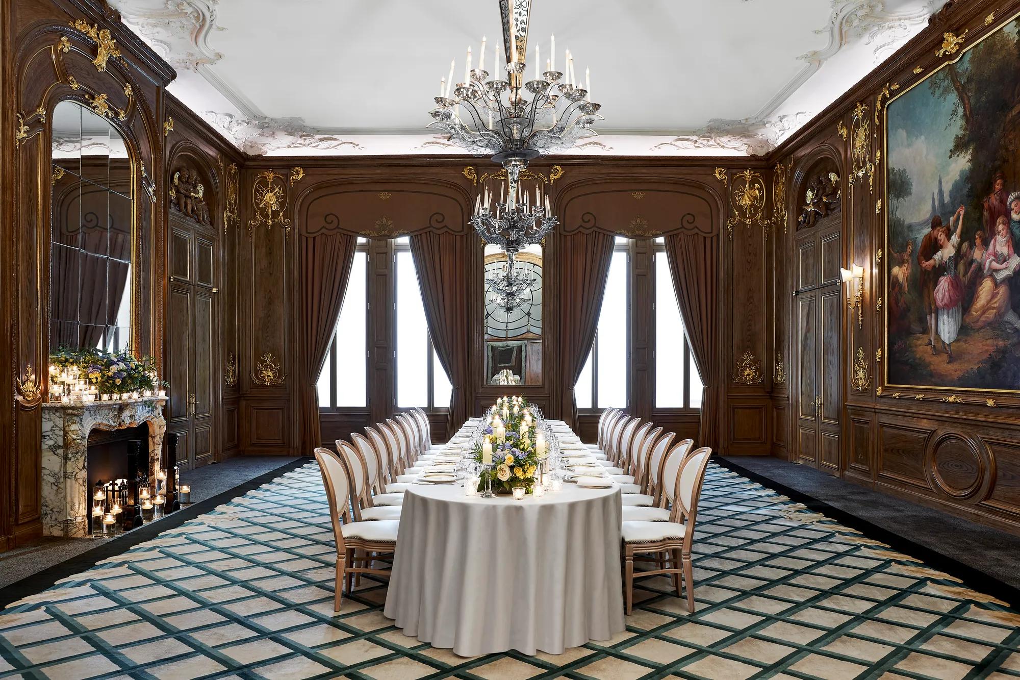 Formal dining room with long table set for event, featuring chandeliers, wood panelling, fireplace, large painting and tall windows with draped curtains.