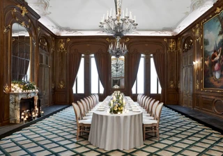 Formal dining room with long table set for event, featuring chandeliers, wood panelling, fireplace, large painting and tall windows with draped curtains.