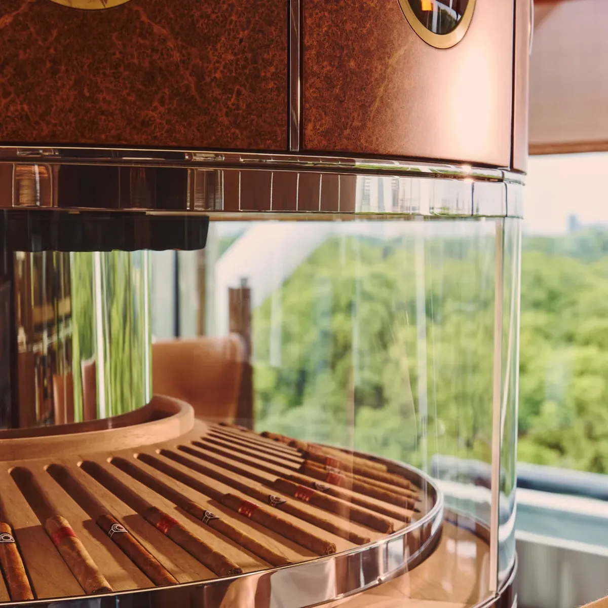A person selecting a cigar from a circular glass humidor with wooden detailing and views of greenery outside.