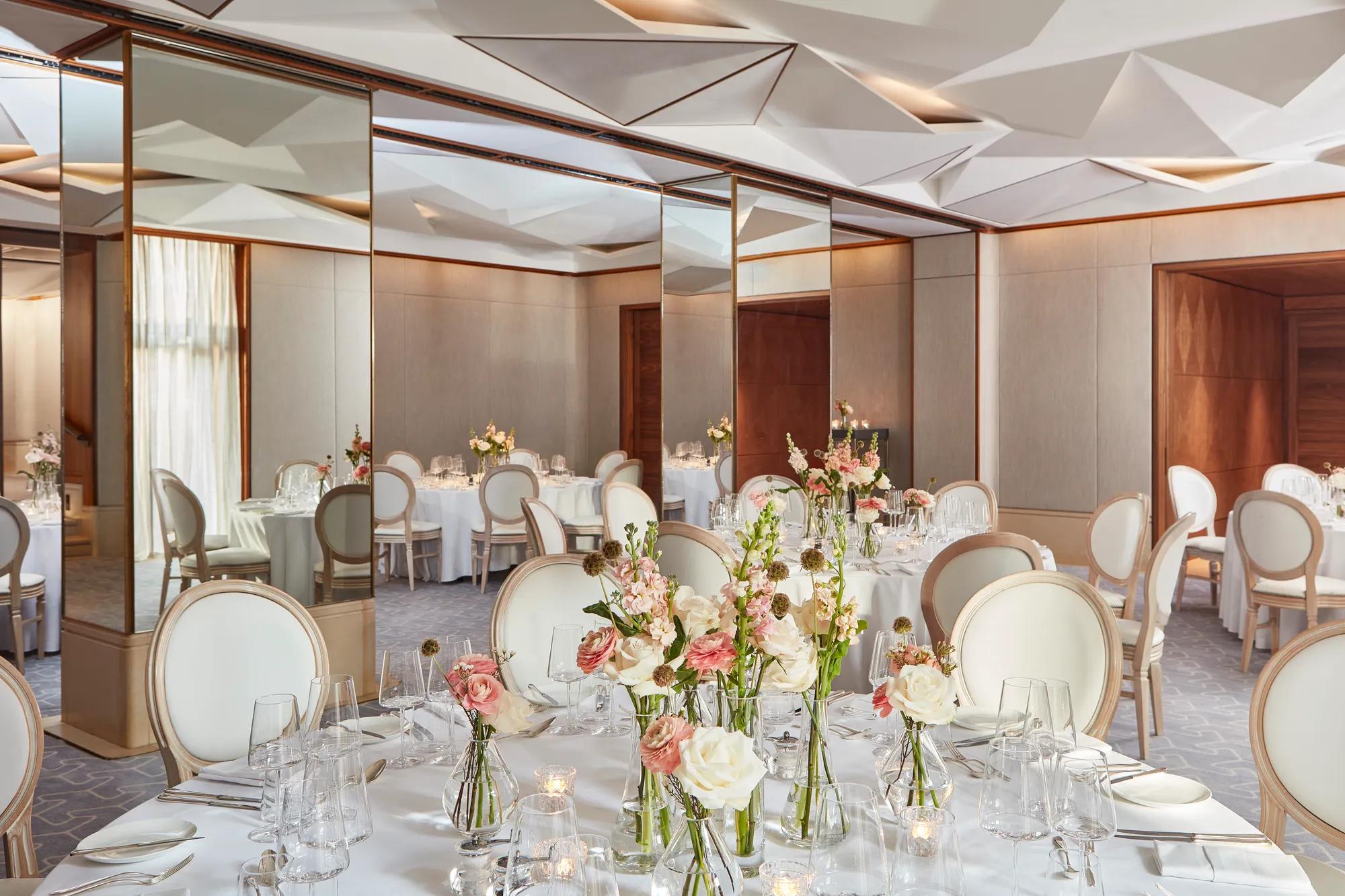 Round tables with white chairs, floral centrepieces, and mirrored walls reflecting soft daylight.