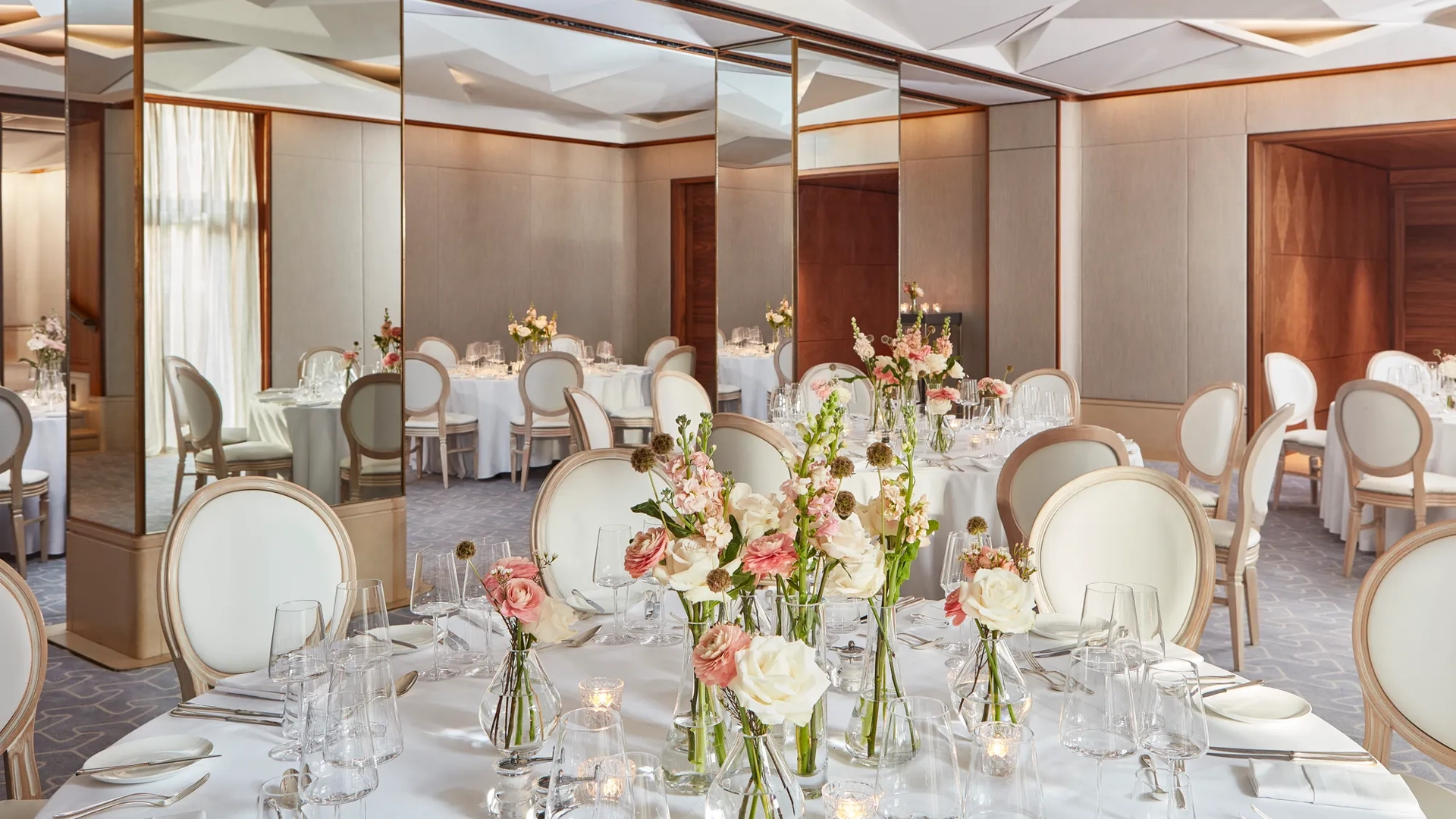 Round tables with white chairs, floral centrepieces, and mirrored walls reflecting soft daylight.