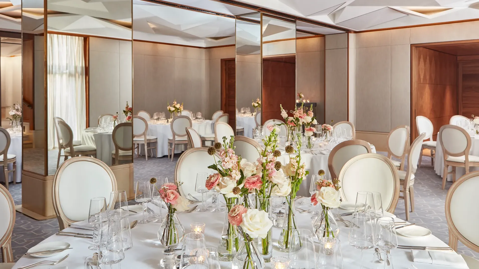 Round tables with white chairs, floral centrepieces, and mirrored walls reflecting soft daylight.