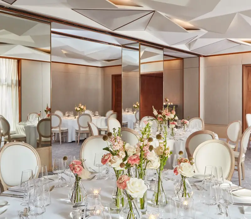 Round tables with white chairs, floral centrepieces, and mirrored walls reflecting soft daylight.