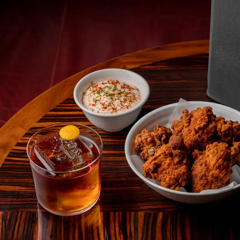 Bowl of golden fried chicken served with a dish of creamy dipping sauce, a dark amber cocktail on ice, and a black Claridge’s Bar menu on a polished wooden table with a small vase of flowers.