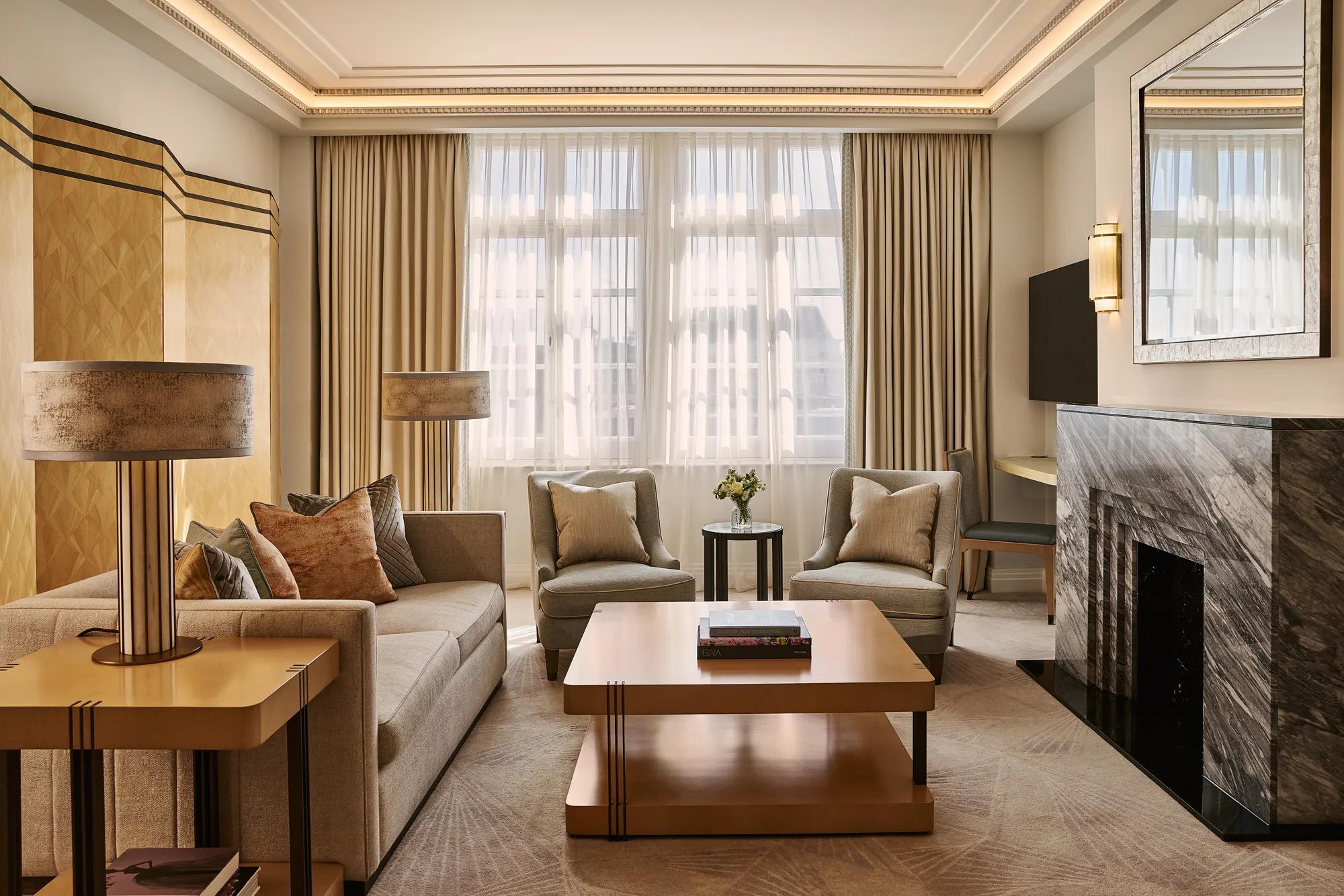 Spacious living room with sofa and armchairs, central coffee table, large window with curtains, and a marble fireplace with mirror.
