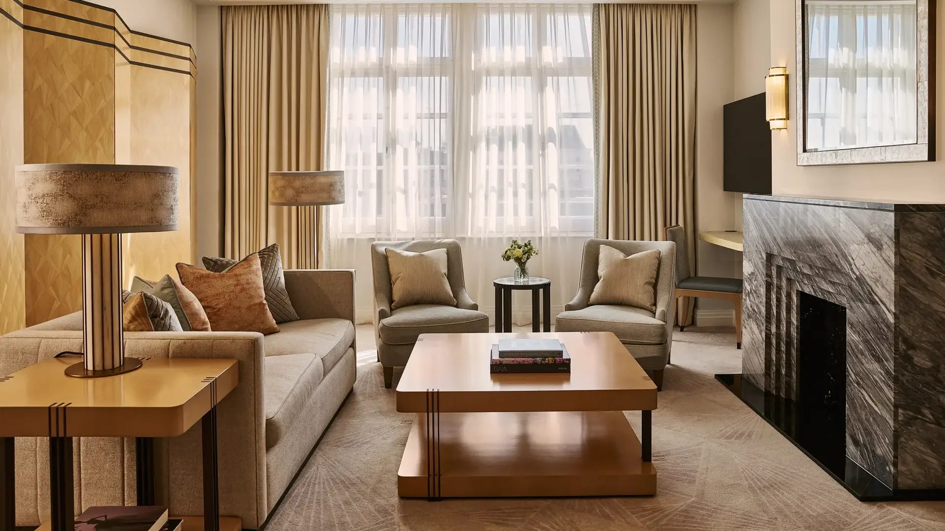 Spacious living room with sofa and armchairs, central coffee table, large window with curtains, and a marble fireplace with mirror.