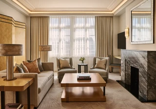 Spacious living room with sofa and armchairs, central coffee table, large window with curtains, and a marble fireplace with mirror.