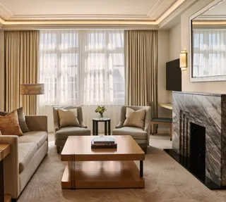 Spacious living room with sofa and armchairs, central coffee table, large window with curtains, and a marble fireplace with mirror.