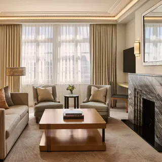 Spacious living room with sofa and armchairs, central coffee table, large window with curtains, and a marble fireplace with mirror.