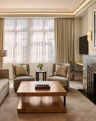 Spacious living room with sofa and armchairs, central coffee table, large window with curtains, and a marble fireplace with mirror.