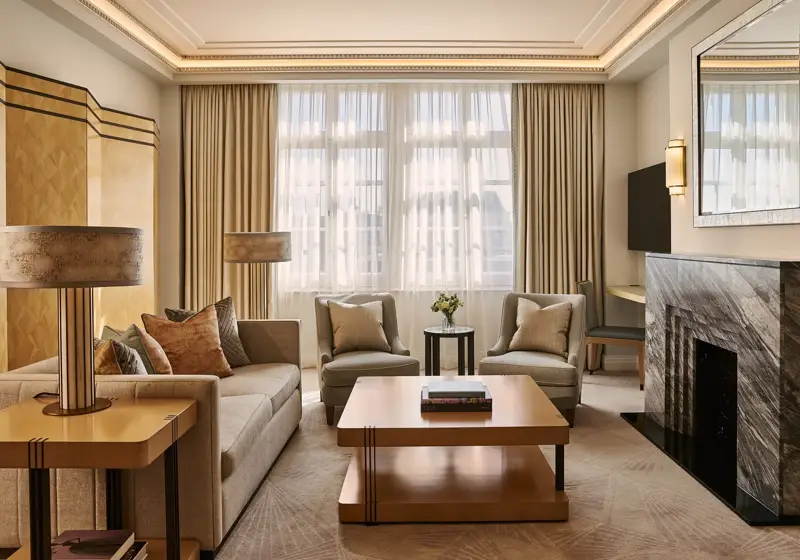 Spacious living room with sofa and armchairs, central coffee table, large window with curtains, and a marble fireplace with mirror.