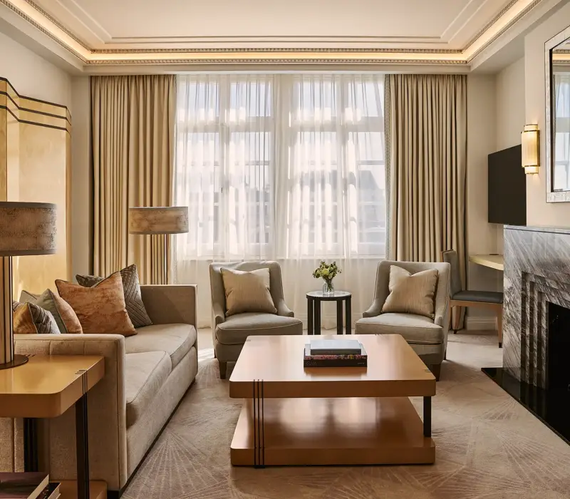 Spacious living room with sofa and armchairs, central coffee table, large window with curtains, and a marble fireplace with mirror.