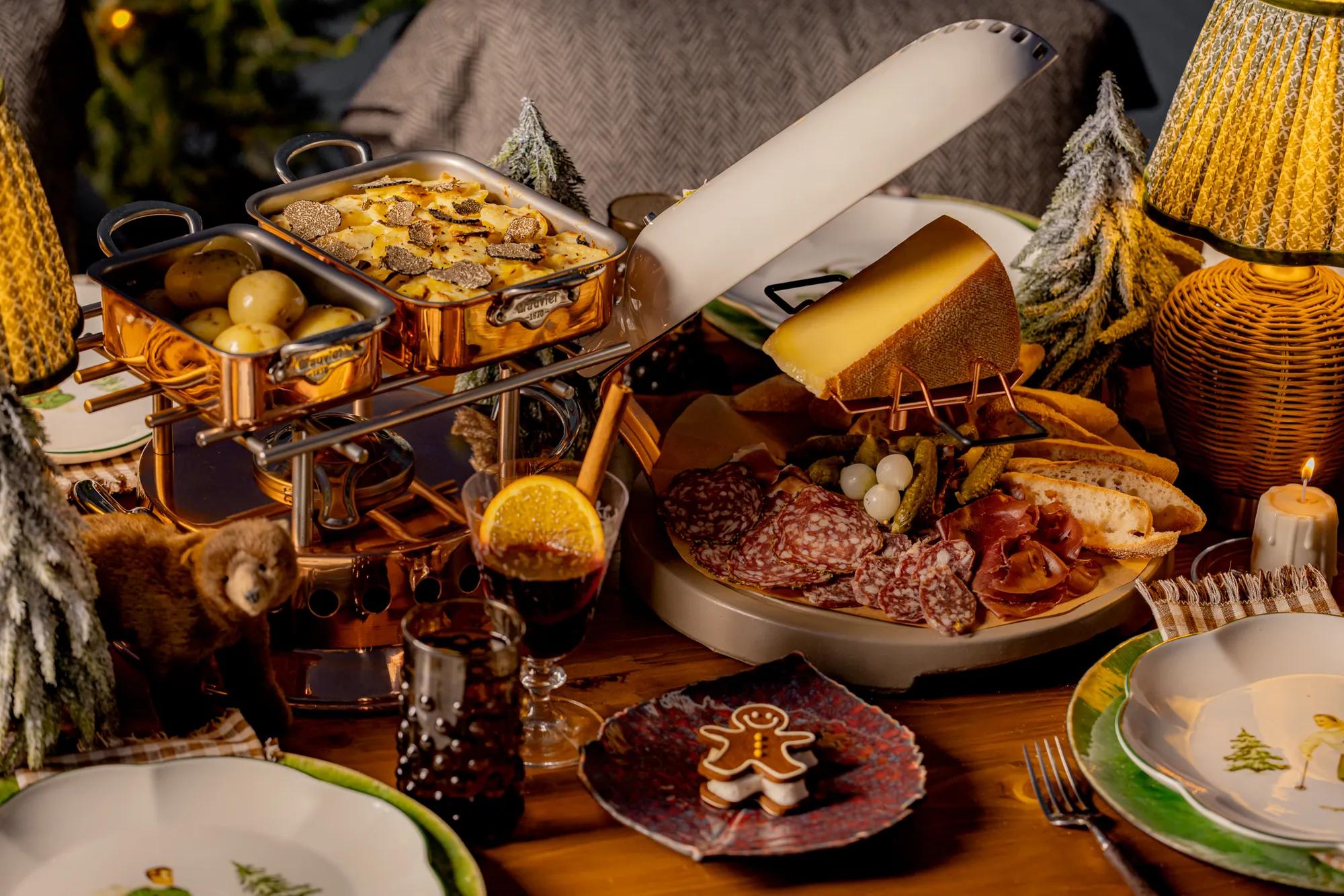Festive dining table with raclette cheese, charcuterie, potatoes and a gingerbread biscuit, surrounded by miniature snowy trees and warm lighting.