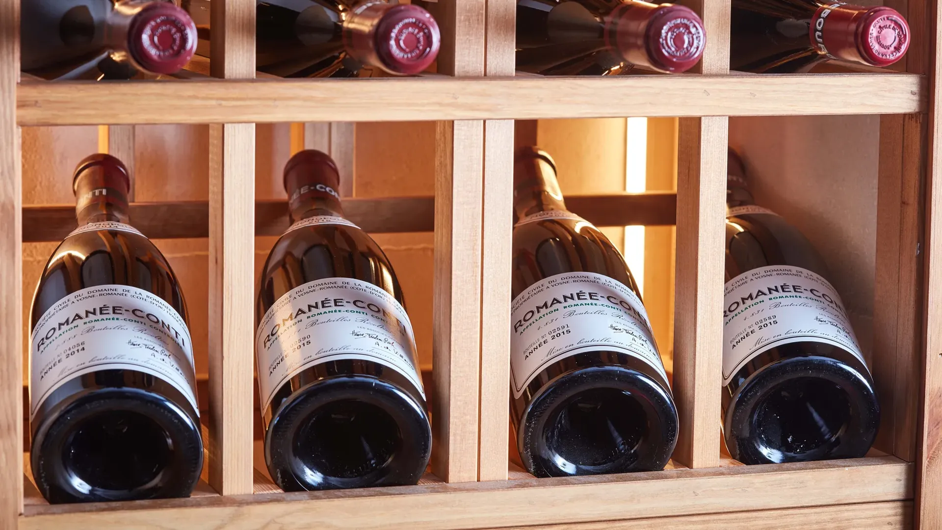 Red wine bottles stored horizontally in a wooden rack, with labels and corks visible under warm lighting.
