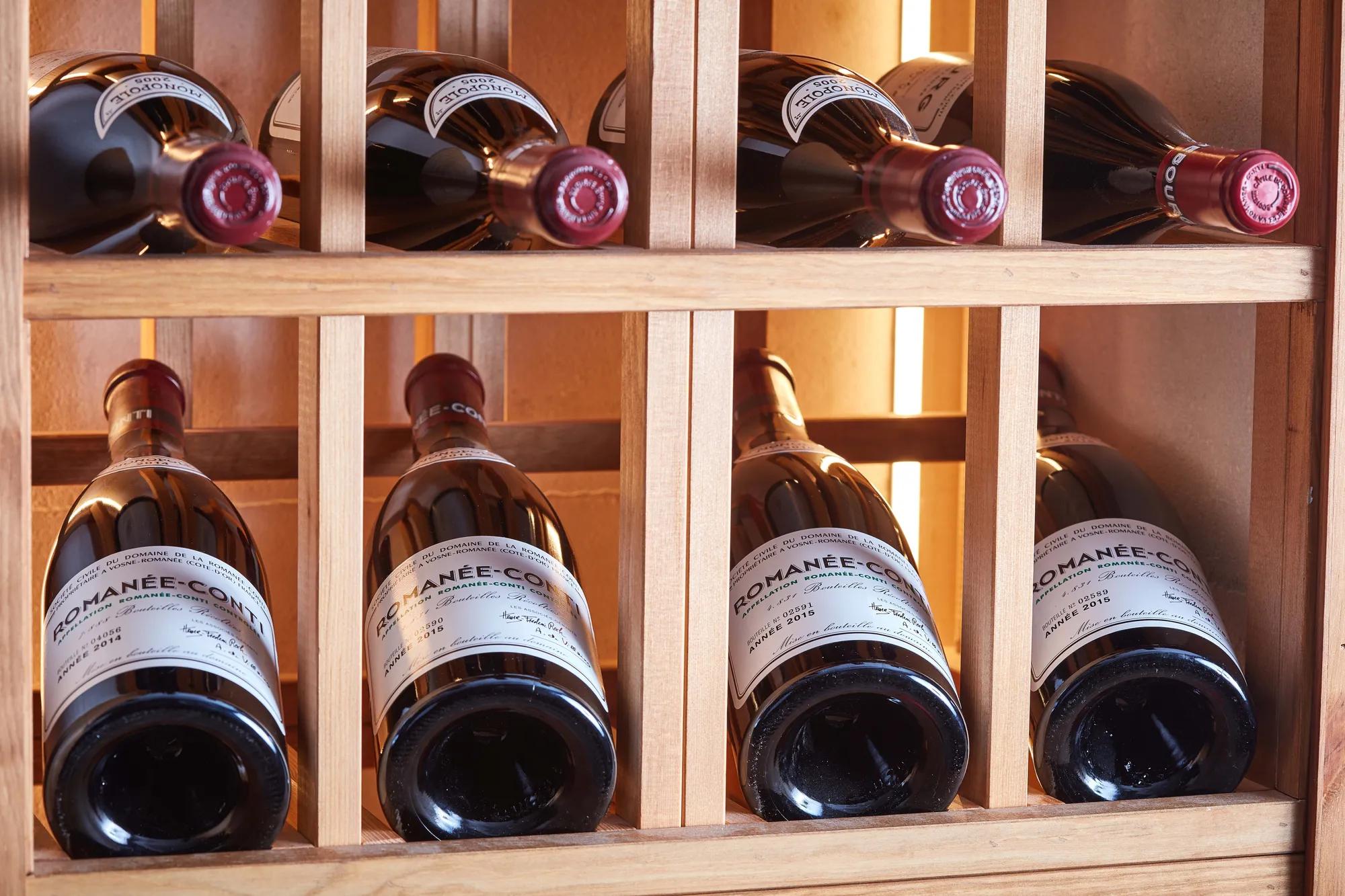 Red wine bottles stored horizontally in a wooden rack, with labels and corks visible under warm lighting.