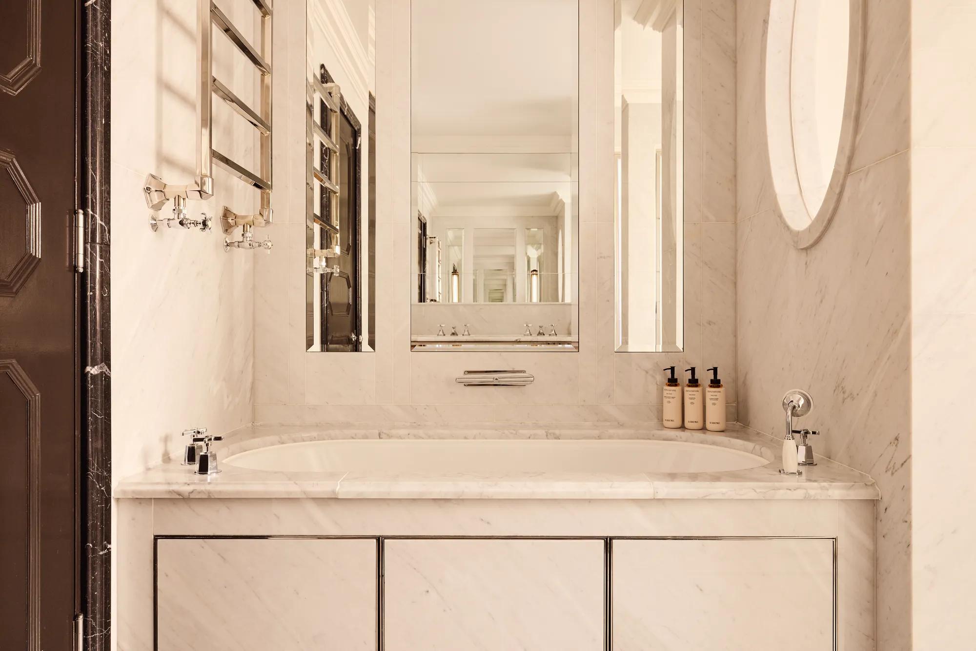 Elegant marble bathroom featuring a built-in bathtub beneath large mirrors, chrome fixtures, and a heated towel rail, with toiletries neatly arranged along the edge.