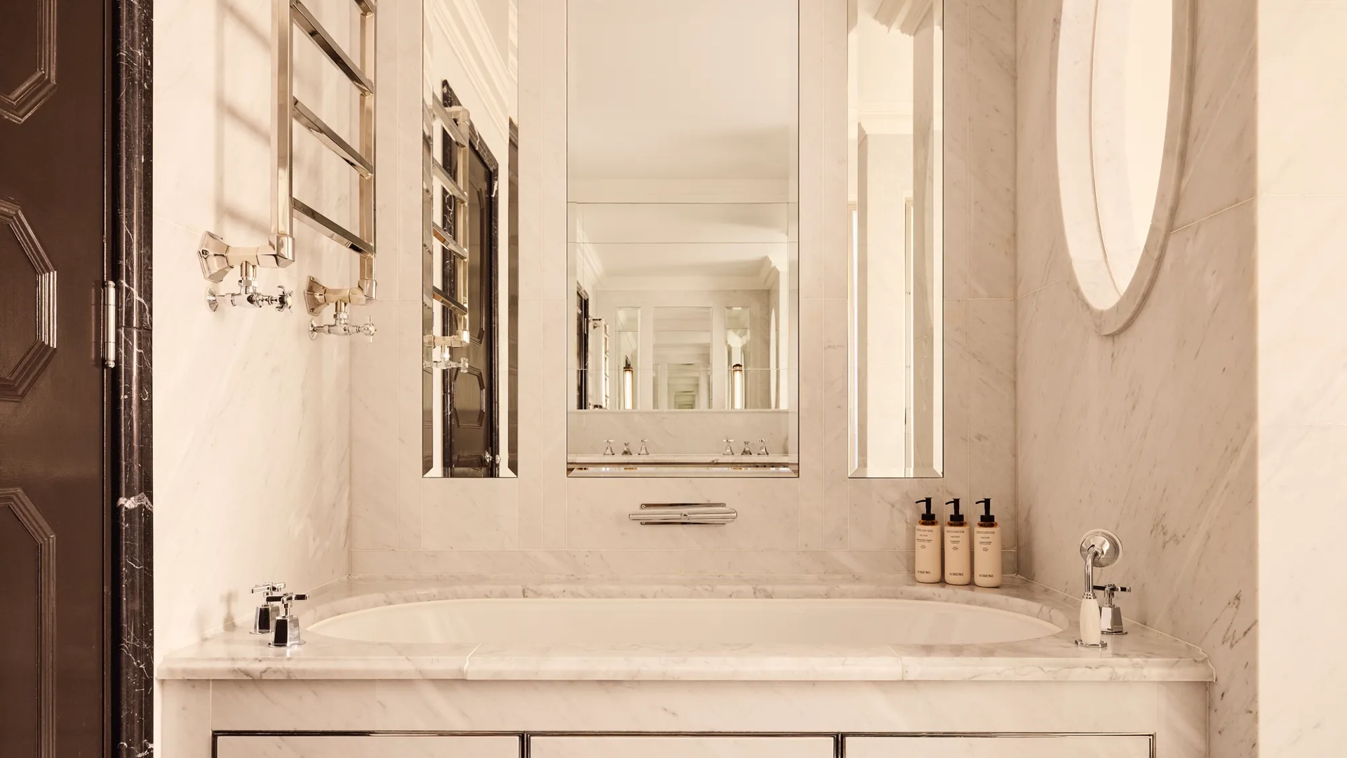 Elegant marble bathroom featuring a built-in bathtub beneath large mirrors, chrome fixtures, and a heated towel rail, with toiletries neatly arranged along the edge.