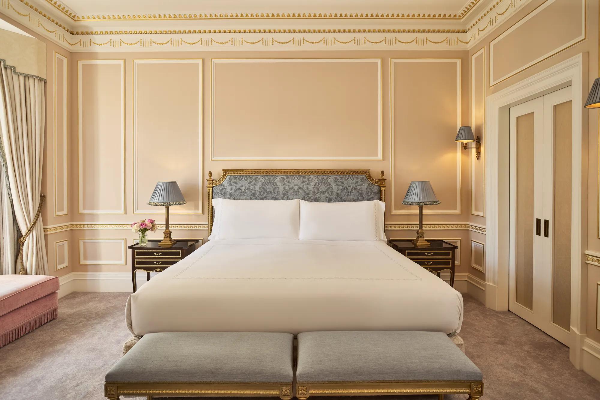 King-size bed with an upholstered headboard, twin bedside tables and benches, set within a classically detailed bedroom with gold-trimmed walls and soft neutral tones.