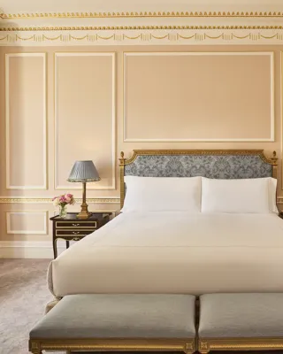 King-size bed with an upholstered headboard, twin bedside tables and benches, set within a classically detailed bedroom with gold-trimmed walls and soft neutral tones.