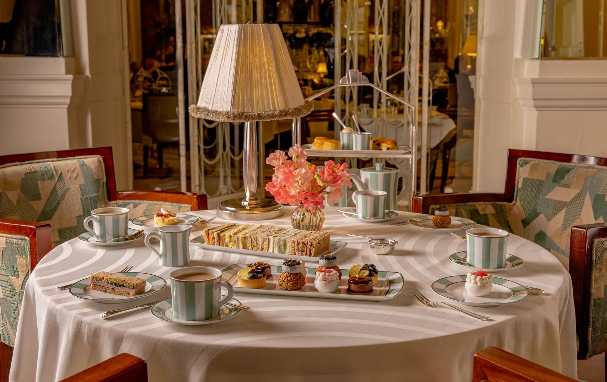 Elegant afternoon tea table set with finger sandwiches, miniature pastries, teacups and pink flowers beneath a fringed table lamp in a refined dining room.