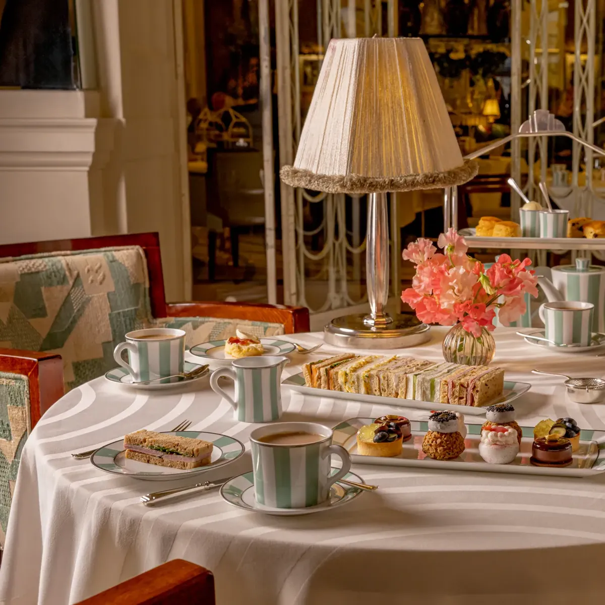 Elegant afternoon tea table set with finger sandwiches, miniature pastries, teacups and pink flowers beneath a fringed table lamp in a refined dining room.