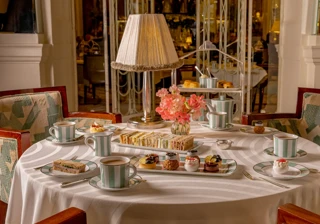 Elegant afternoon tea table set with finger sandwiches, miniature pastries, teacups and pink flowers beneath a fringed table lamp in a refined dining room.
